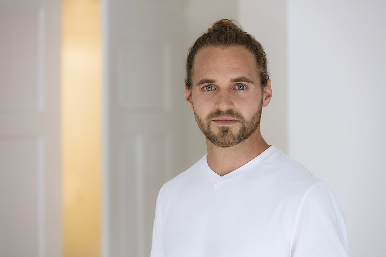 Ein junger Mann mit blondem Haar, Bart und blauen Augen trägt ein weißes T-Shirt, steht vor einem hellen Hintergrund.