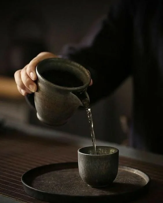 Person gießt heißen Kaffee aus einer dunklen Tasse in eine kleinere Tasse auf einem runden Tablett.