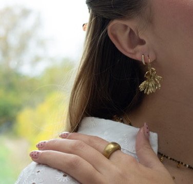 Nærbillede af en kvinde med ørepynt og ring, der rører ved hendes skulder, udendørs med grøn baggrund.