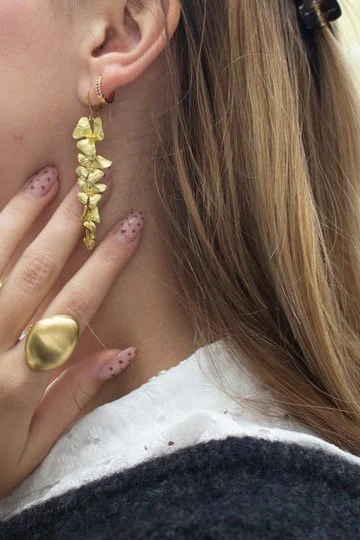 Nærbillede af en kvindes øre med store gule øreringe, en hånd med en stor gul ring og pletmønstrede negle tæt på øret.