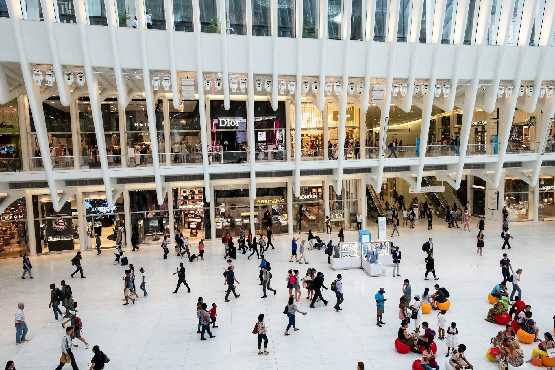 Des gens marchent et font du shopping dans un grand centre commercial moderne à plusieurs niveaux, avec des magasins comme Dior et Breitling visibles.