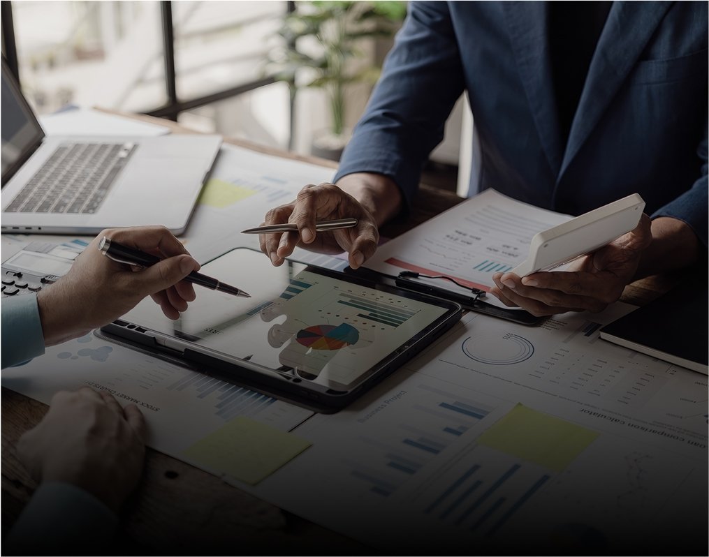 Réunion d'affaires avec deux personnes discutant de données financières, à l'aide d'une tablette avec des graphiques, d'un ordinateur portable, de rapports imprimés et d'un smartphone sur une table en bois.