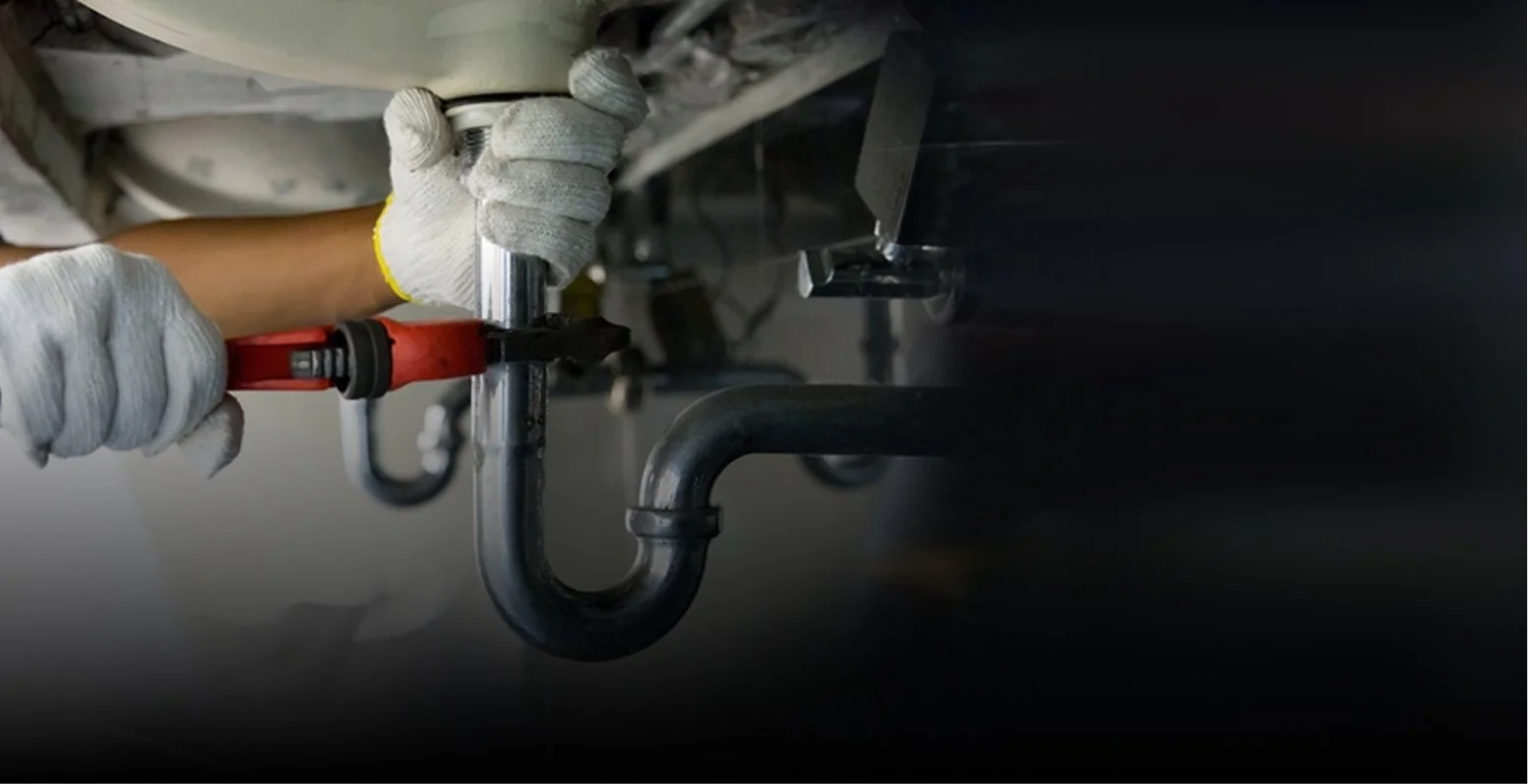 Un ouvrier portant des gants blancs resserre un tuyau métallique sous un évier ou un appareil de plomberie similaire à l'aide d'une clé rouge.