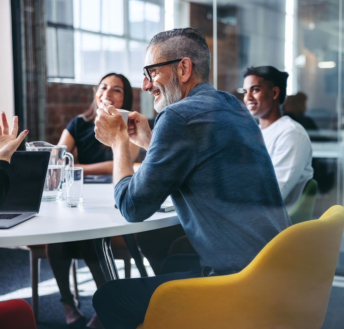 Groupe de quatre personnes assises autour d'une table dans un bureau moderne, riant et discutant. Un homme avec des lunettes et une barbe tient une tasse, buvant peut-être du café, tandis que les autres écoutent et sourient.