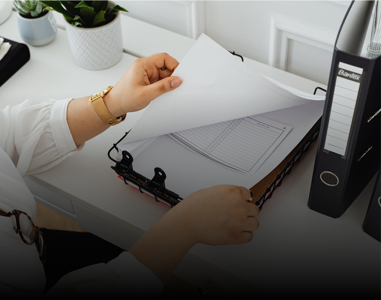 Une personne avec une montre en or tournant les pages d'un classeur sur un bureau avec des plantes en pot et des fournitures de bureau à proximité.