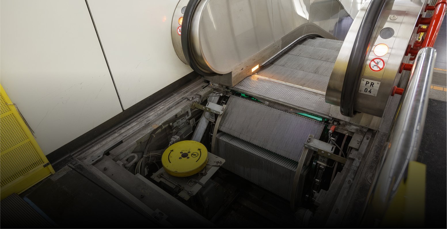 Gros plan sur un carrousel à bagages d'aéroport avec une courroie métallique et un couvercle de sécurité jaune avec un autocollant représentant un visage heureux. La zone semble être en cours d'entretien ou de réparation, certains revêtements ayant été soulevés pour révéler les engrenages et les mécanismes sous-jacents.