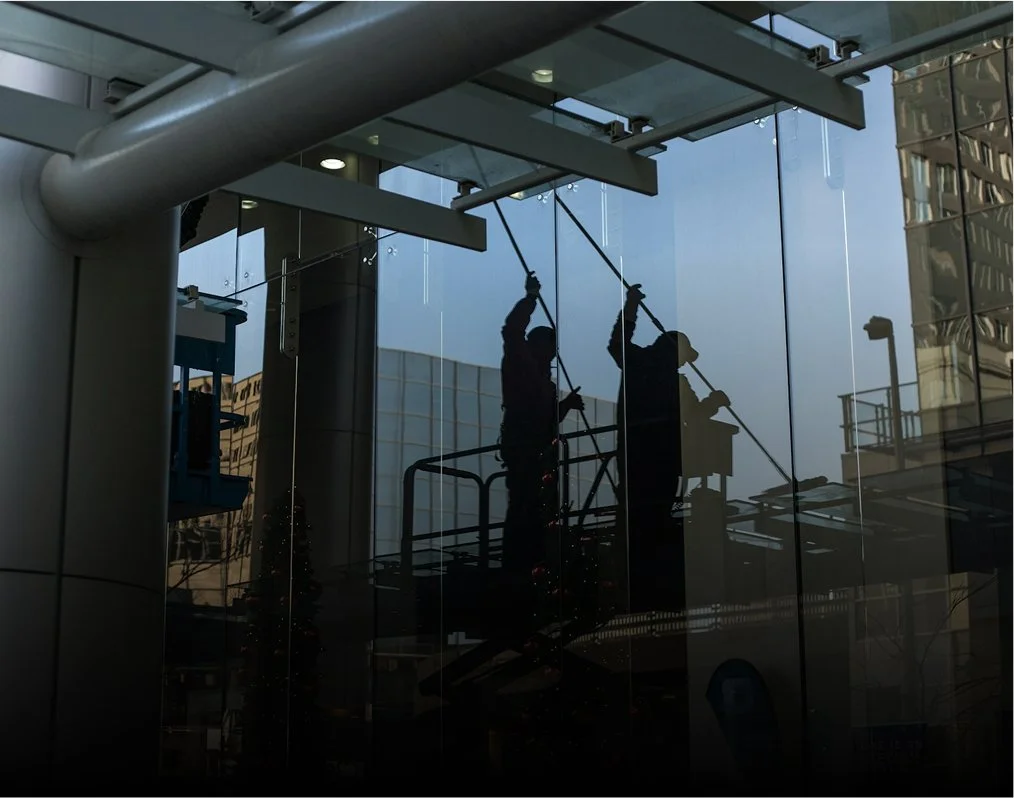 Fenêtre en verre réfléchissant montrant deux travailleurs nettoyant l'extérieur, avec des bâtiments urbains se reflétant dans la vitre.