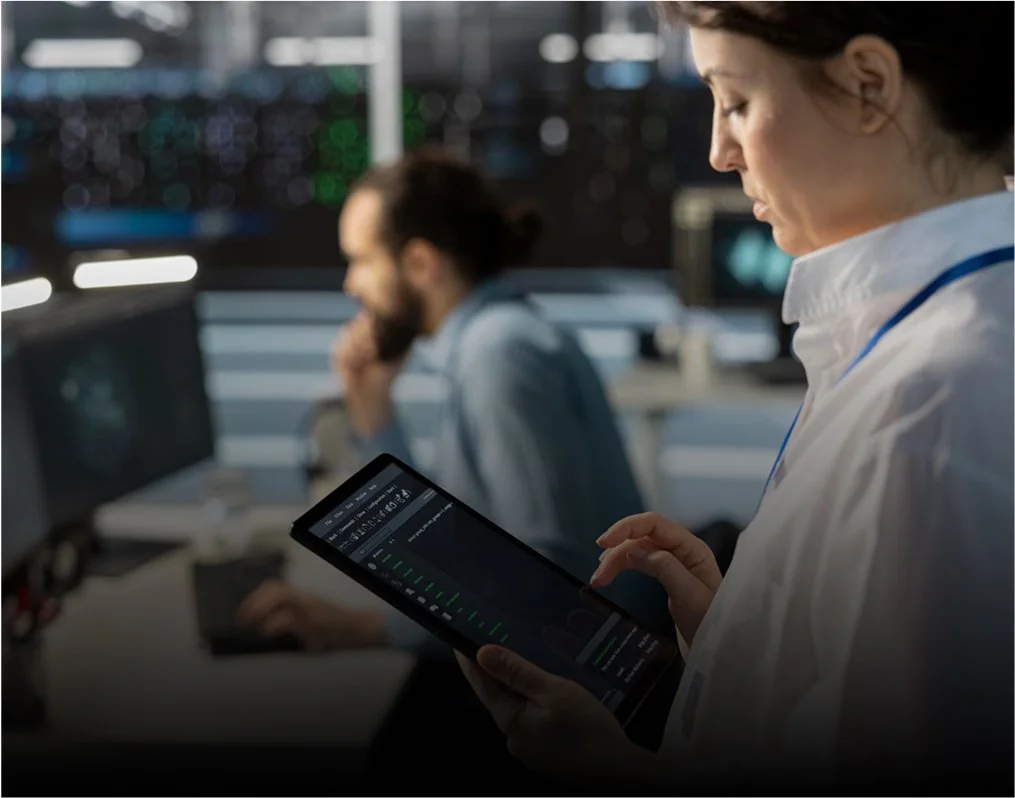 Una mujer con bata blanca de laboratorio utiliza una tableta en una sala de control de alta tecnología, con un hombre al fondo trabajando con un ordenador.