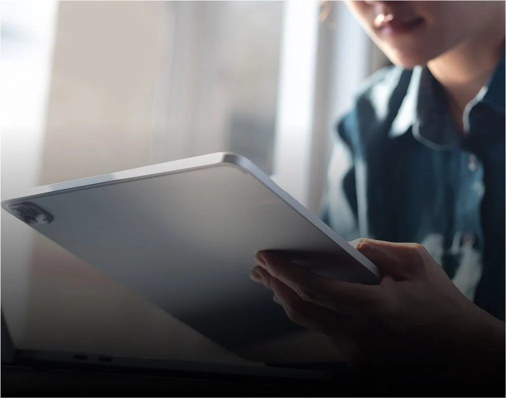 Personne tenant une tablette près d'un ordinateur portable, visage partiellement visible, dans une pièce bien éclairée.