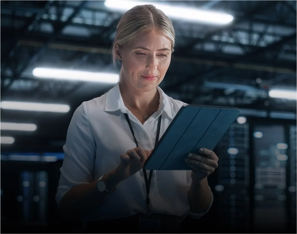 Une femme aux cheveux blonds, vêtue d'une chemise blanche et d'un cordon noir, debout dans un centre de données ou une salle de serveurs, regardant une tablette et interagissant avec elle.