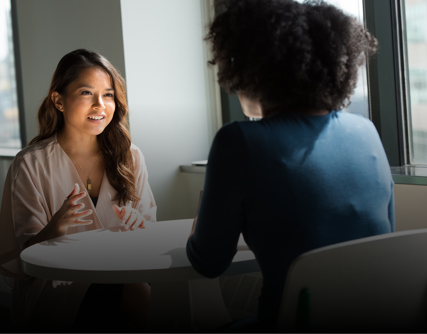 Dos mujeres conversan en una mesa cerca de una ventana, una habla y gesticula, la otra escucha.