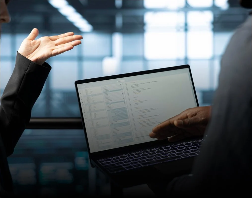 Dos personas en traje de negocios mantienen una discusión en una oficina moderna, una de ellas gesticulando y la otra utilizando un ordenador portátil en el que se muestran códigos o datos.