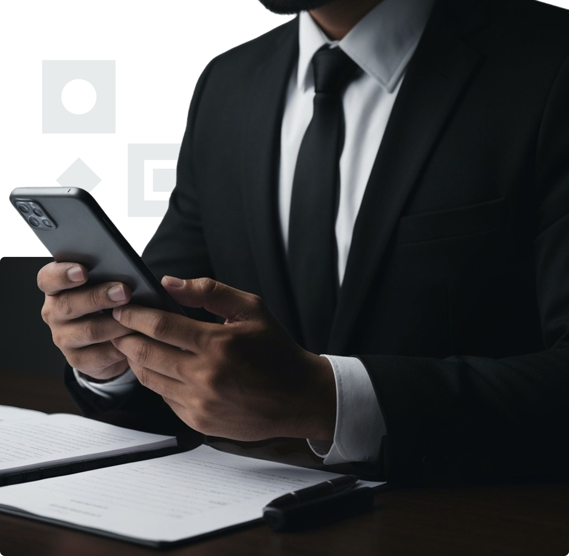 Un homme en costume noir, chemise blanche et cravate noire tenant un smartphone. Un cahier ouvert et un stylo sont posés sur un bureau devant lui.