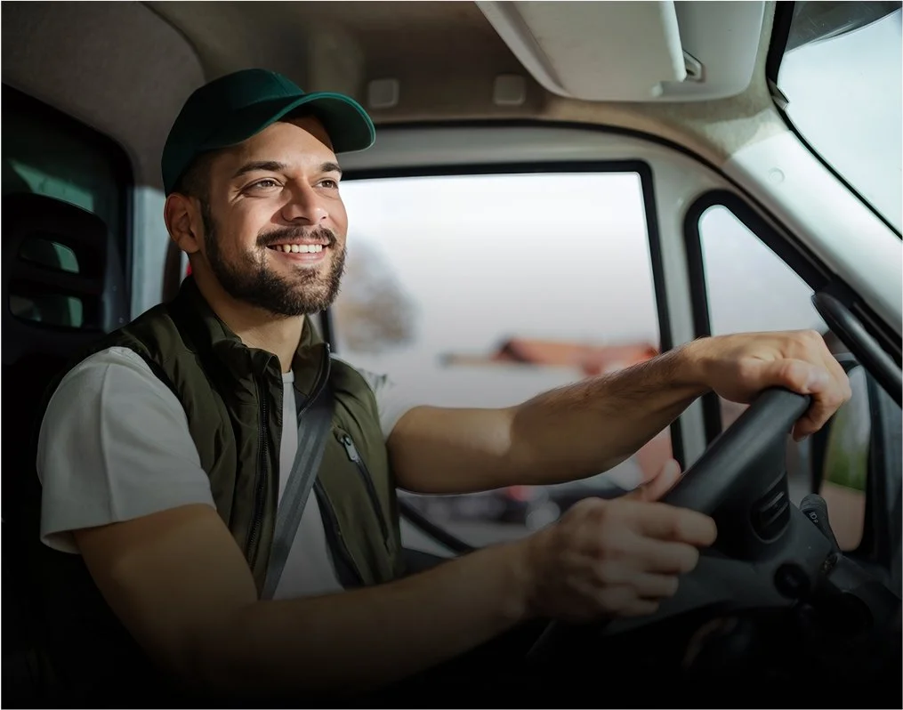 Homme souriant conduisant un camion, portant une casquette et un gilet à l'intérieur du véhicule.