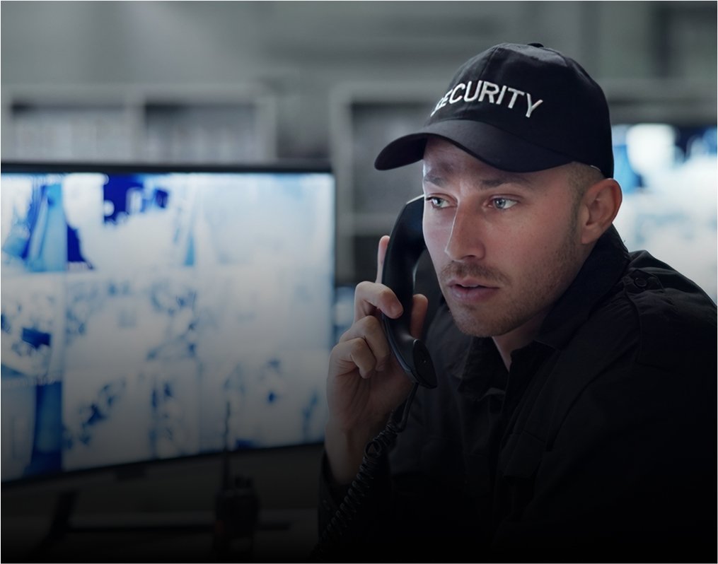 Agent de sécurité avec une casquette portant l'inscription "SECURITY" parlant sur un téléphone fixe et surveillant plusieurs écrans.
