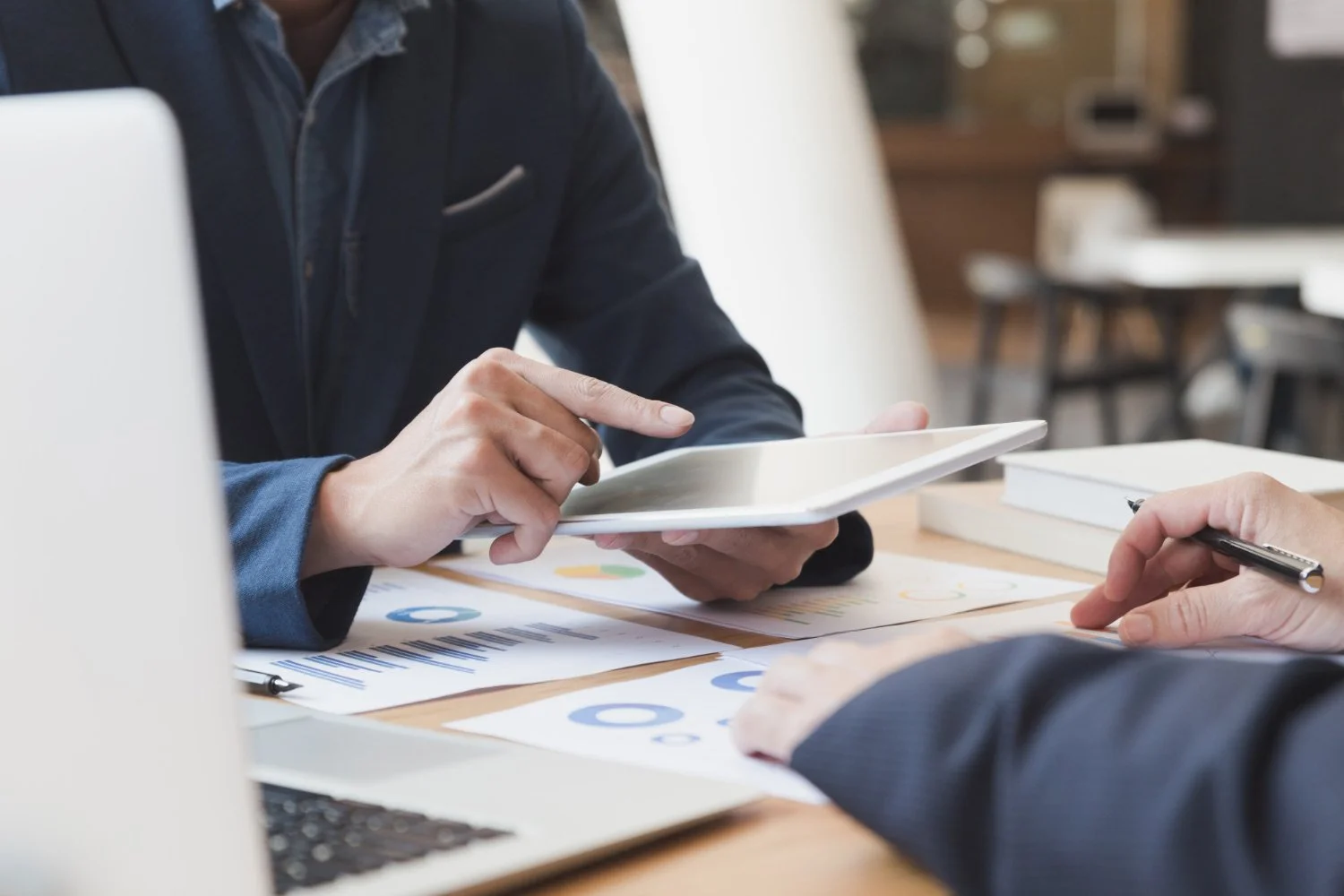 Deux personnes en tenue de travail discutent de documents et d'une tablette à un bureau de travail, avec des tableaux et des graphiques sur les papiers.