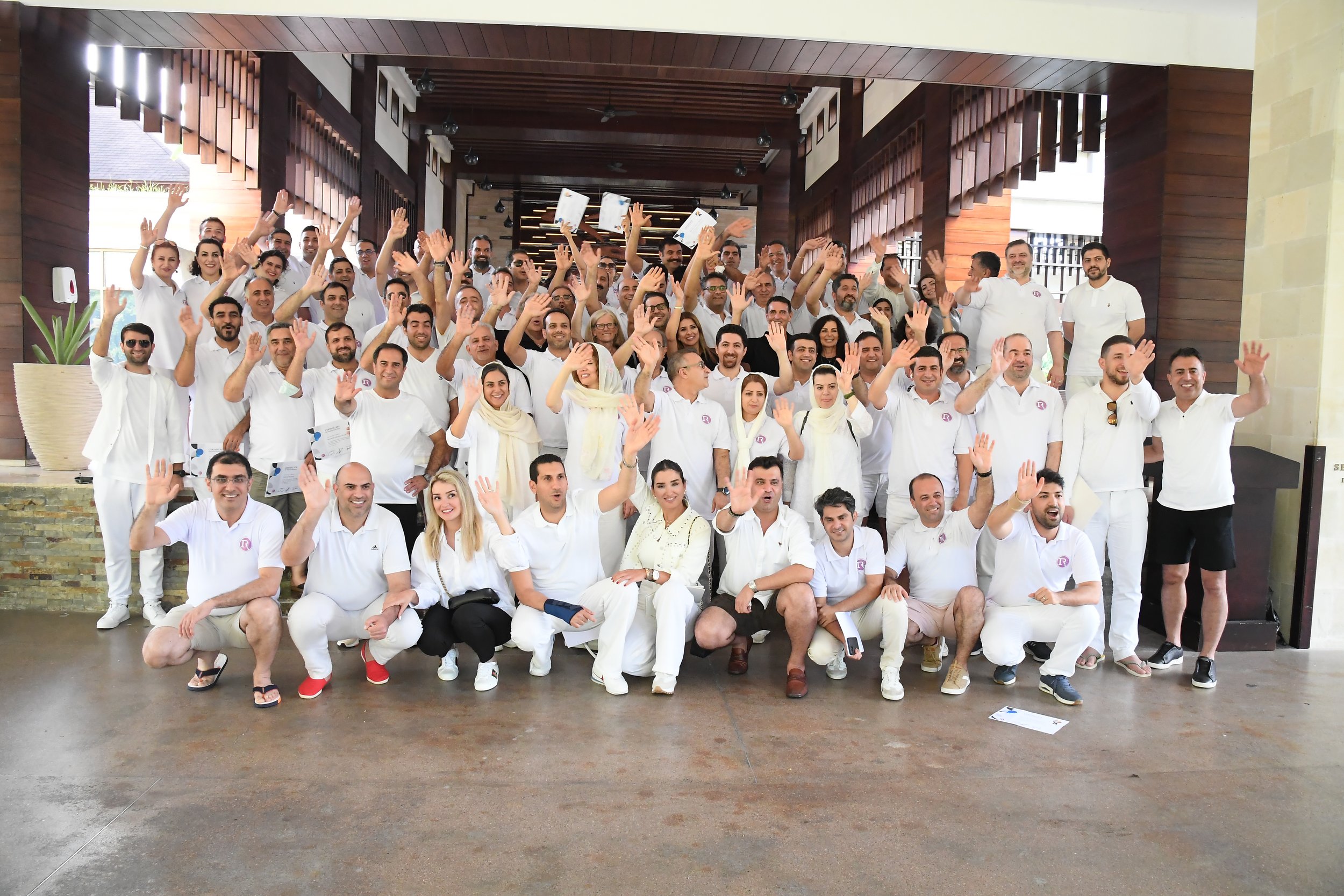 A large group of people dressed in white clothing posing together indoors, many waving and smiling at the camera.