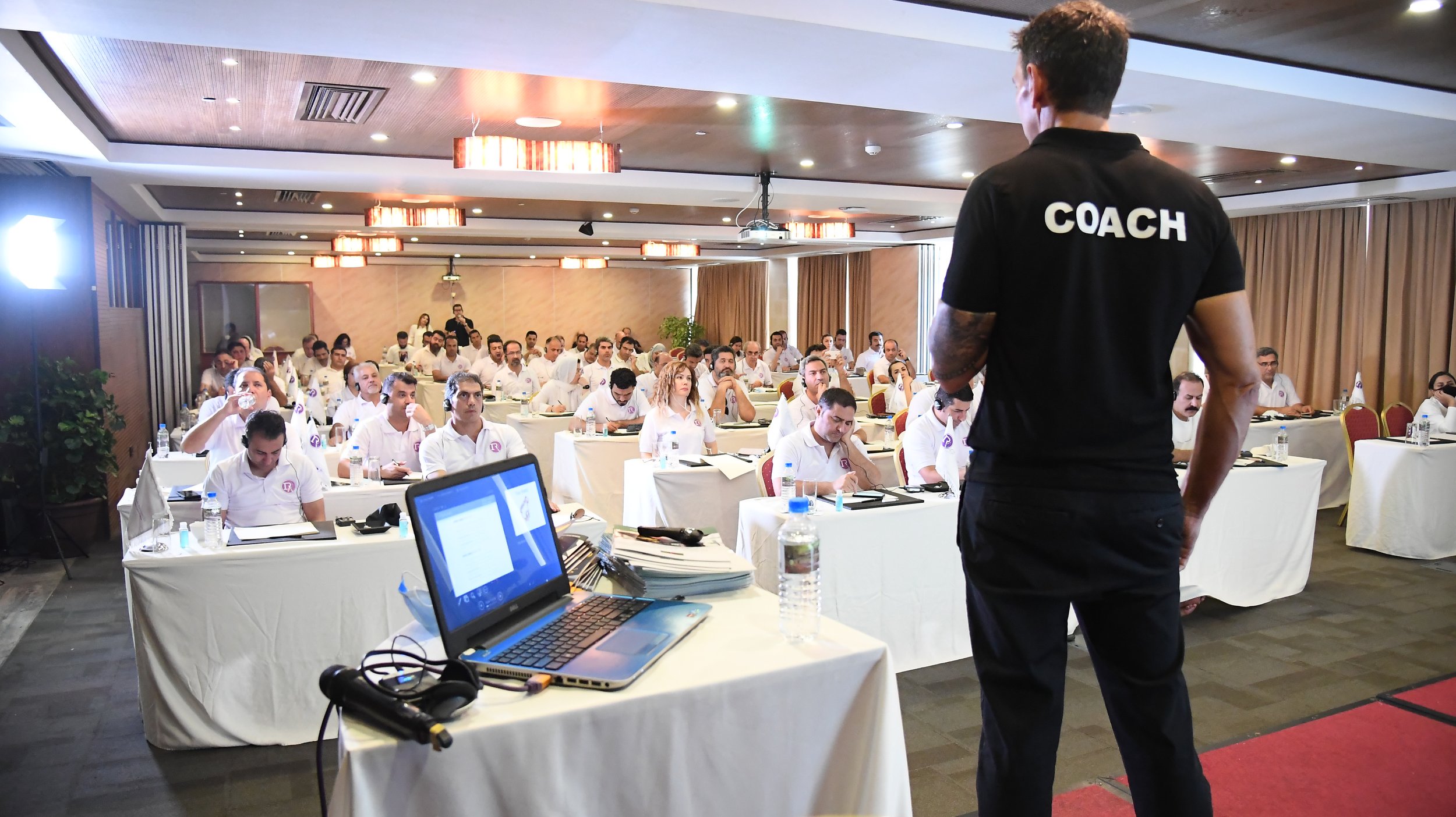 A man with a 'COACH' shirt standing in front of a large audience in a conference room, with attendees sitting at tables and listening.