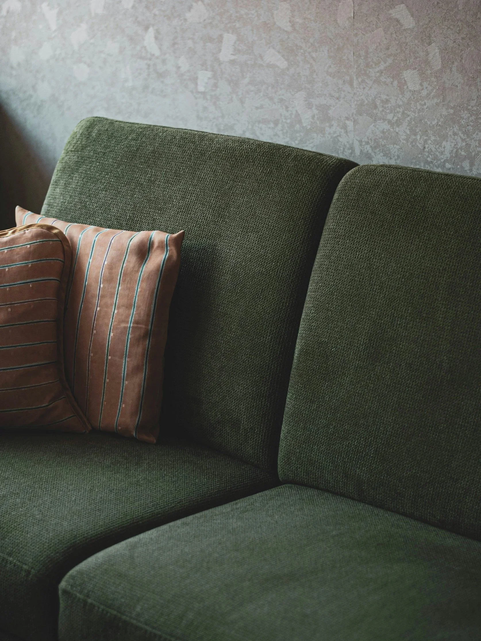 Close-up of a green fabric sofa with a brown striped pillow against a textured gray wall.