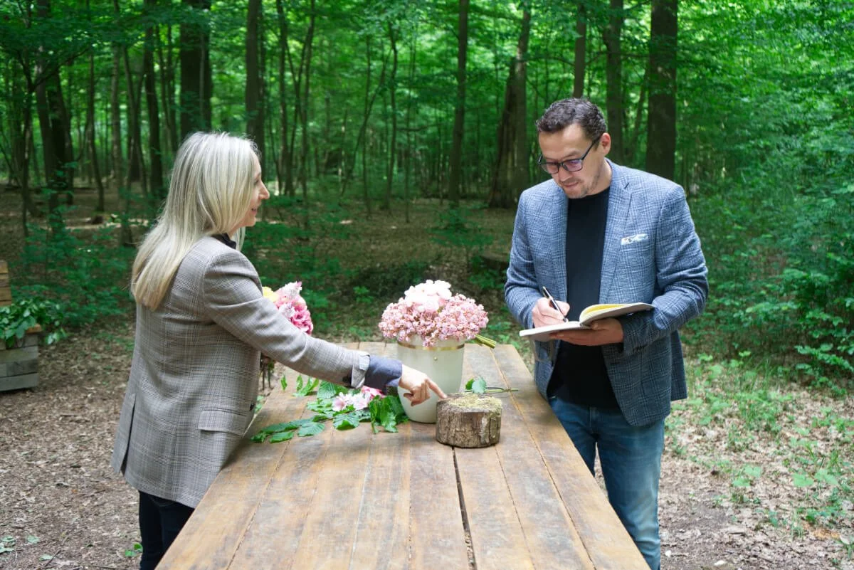 Anke Bachmann und Jens stehen an einem Holztisch im Friedwald und besprechen florale Details; Anke Bach zeigt auf ein Holzstück, während ein Mann Notizen macht – Vorbereitung einer Naturbestattung mit persönlicher Abstimmung bei Veerman Bestattungen.