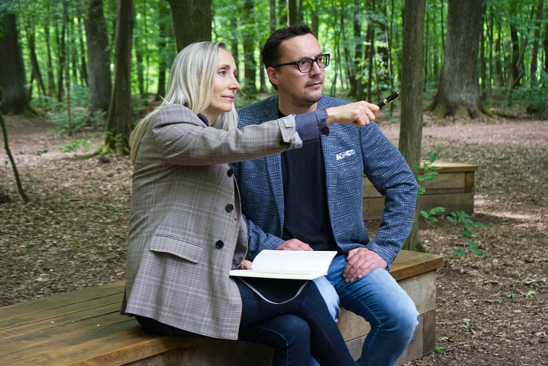 Anke Bachmann und Jens Hasert sitzen auf einer Holzbank im Friedwald. Sie zeigt mit einem Stift in die Ferne, während er aufmerksam schaut; auf ihrem Schoß liegt ein geöffnetes Notizbuch – Szene einer Beratung oder Planung im naturnahen Kontext.