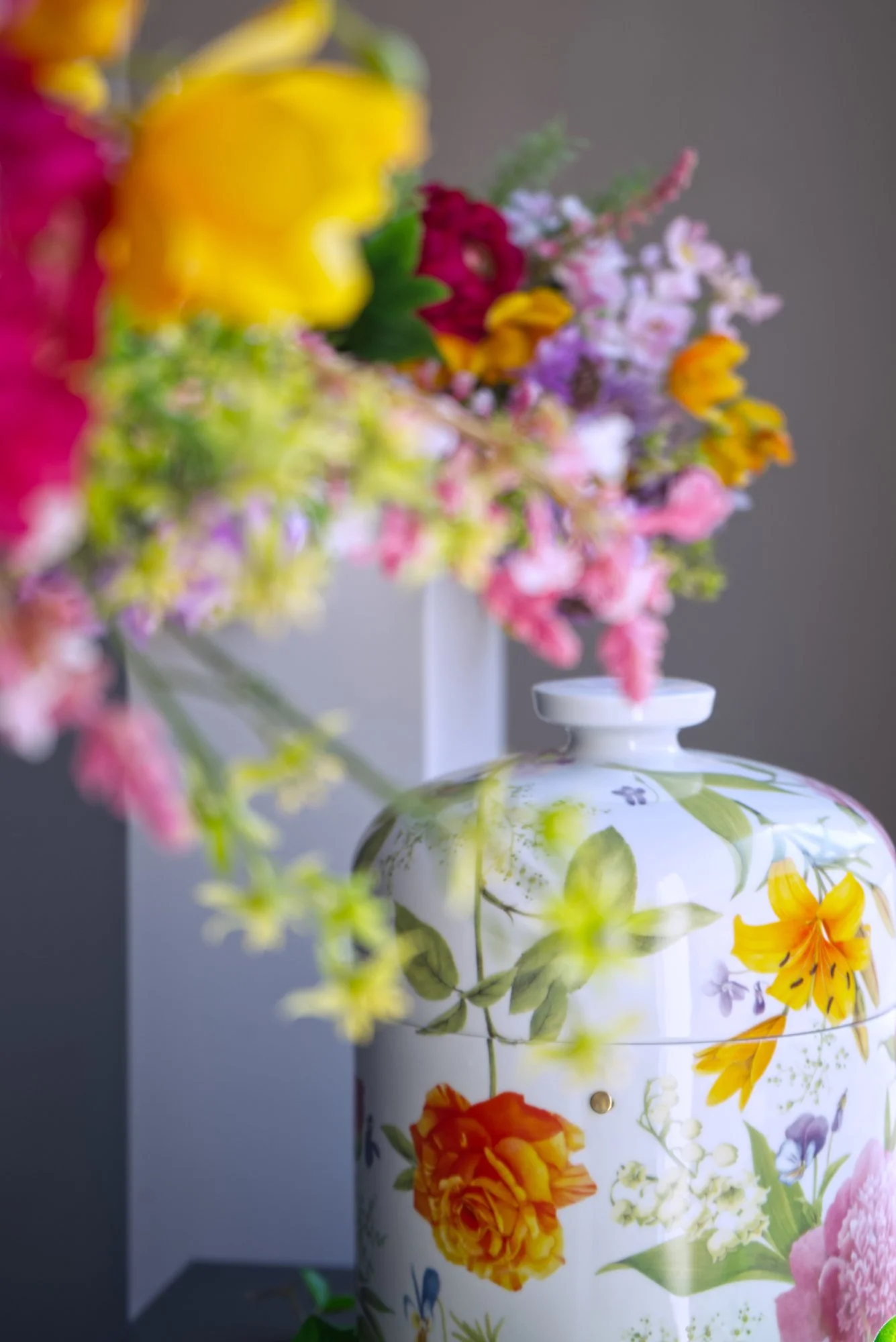 Stillleben mit einer weißen Urne im Vasenstil mit bunten Blumen als Muster. Im Vordergrund ragen unscharf bunte Blumen ins Bild.