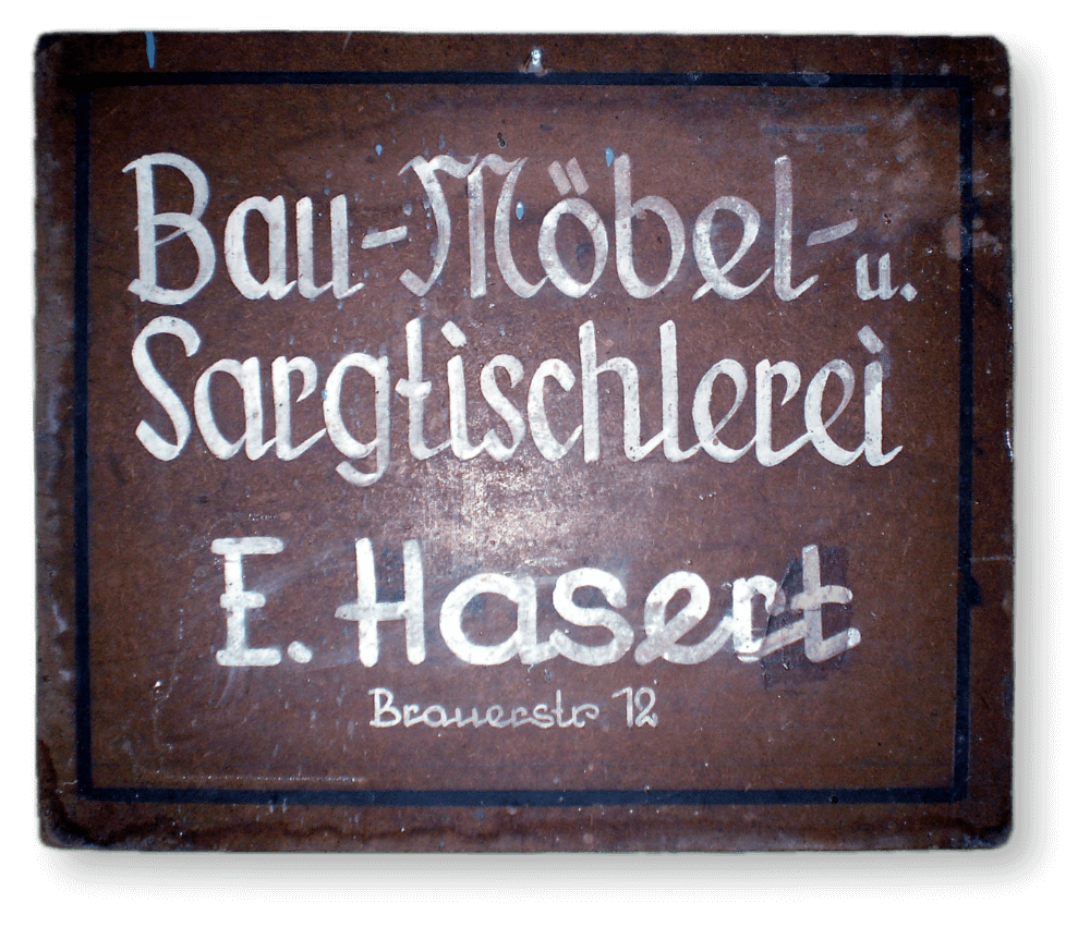Altes, rechteckiges Firmenschild aus Holz mit der Aufschrift „Bau-Möbel- u. Sargtischlerei E. Hasert, Brauerstr. 12“ in weißer, handgemalter Frakturschrift auf braunem Hintergrund.