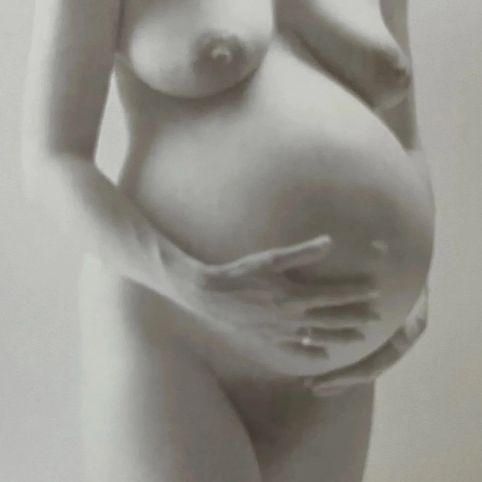 A black and white photograph of a nude woman with breasts exposed, one hand on her stomach, and the other hand outlet cover visible on the wall.