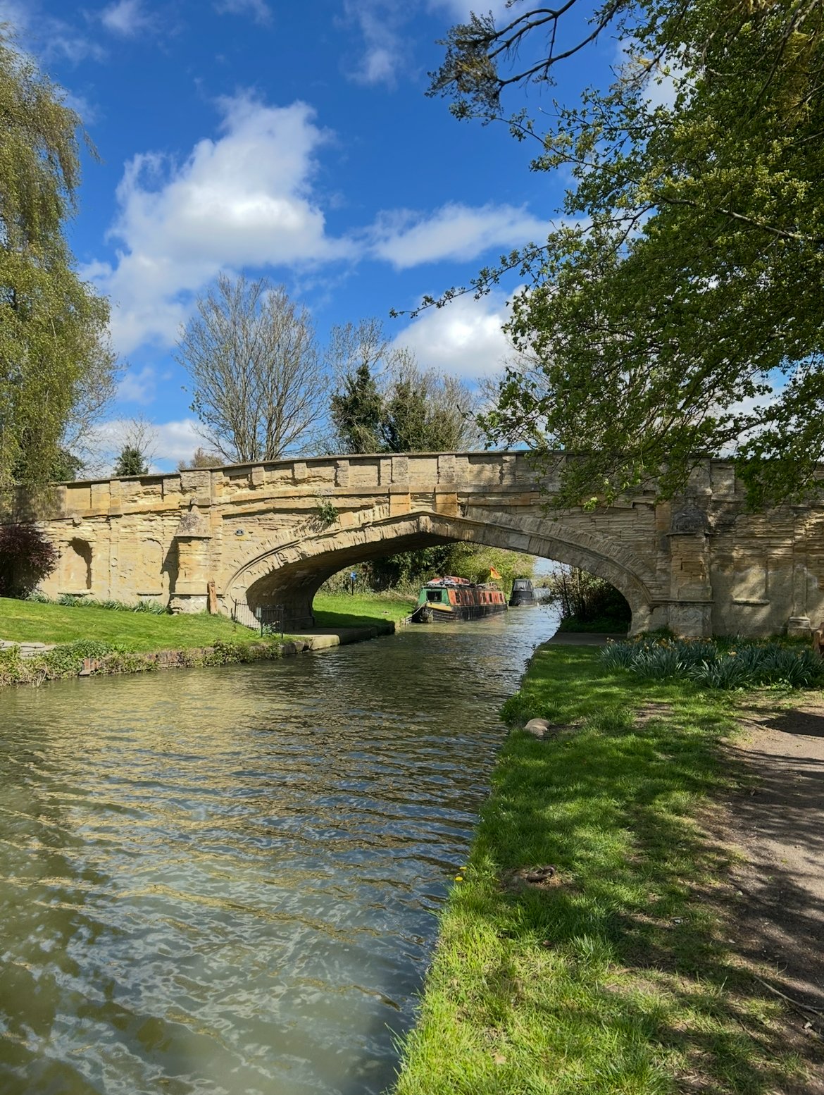 Stone bridge over a canal with a narrowboat passing underneath, surrounded by grassy banks and trees, under a blue sky with clouds.