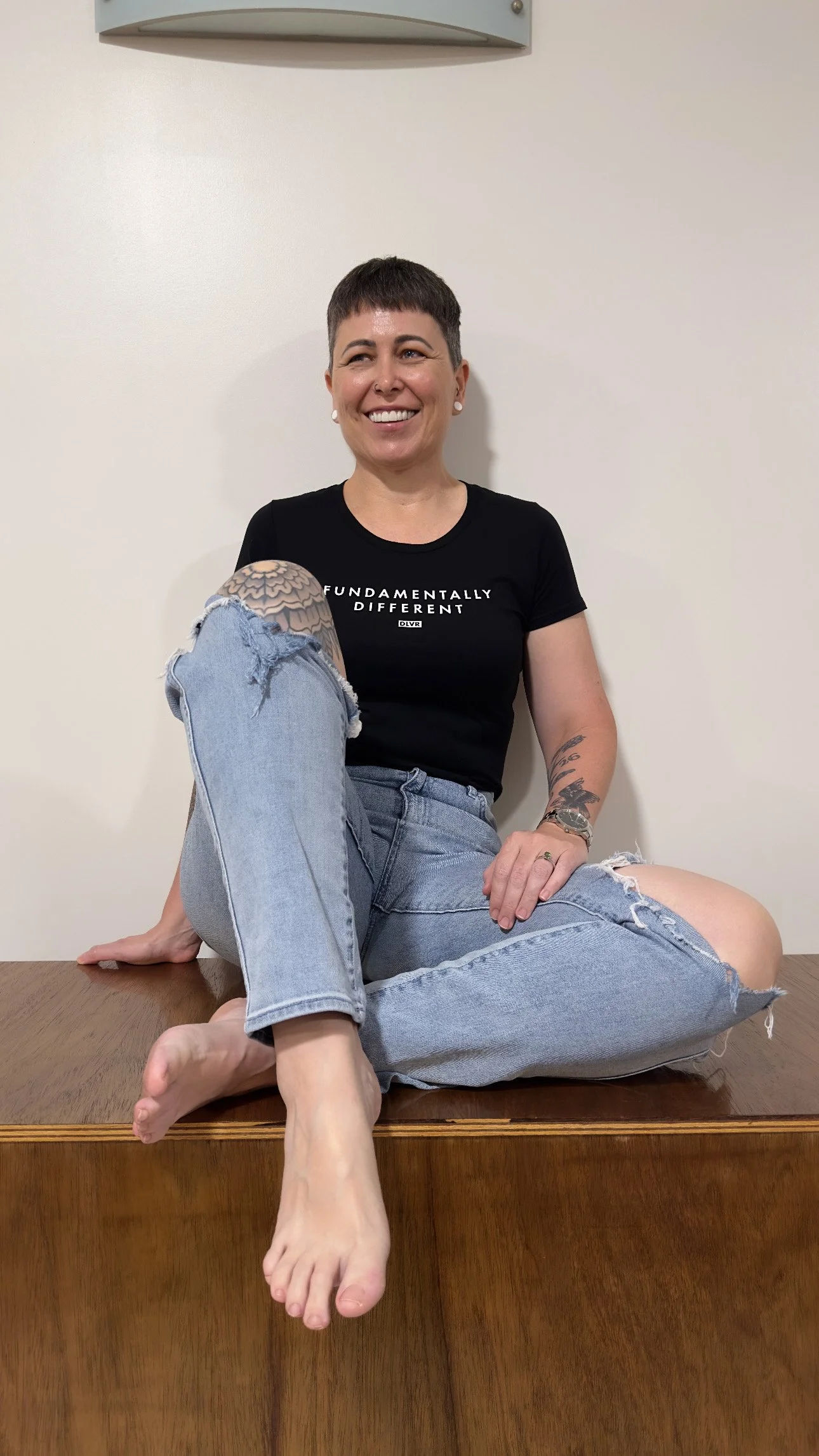 A woman with short dark hair and tattoos sitting on a wooden surface, smiling, wearing a black T-shirt and ripped jeans.