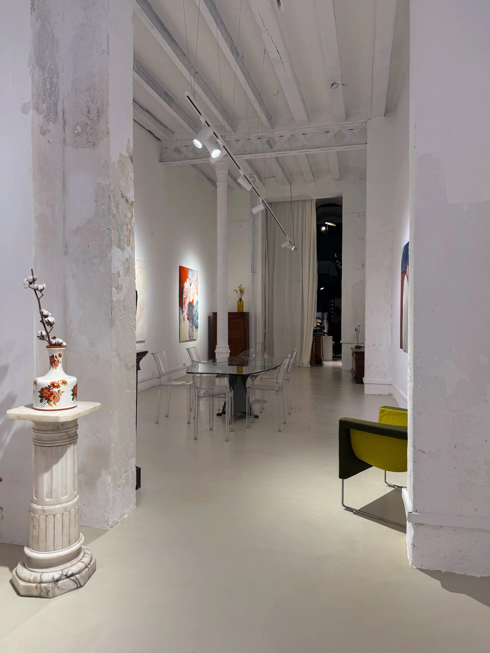 View of Matiz Gallery interior featuring textured white walls, marble pedestal with ceramic vase, and glass dining table under exposed beams.