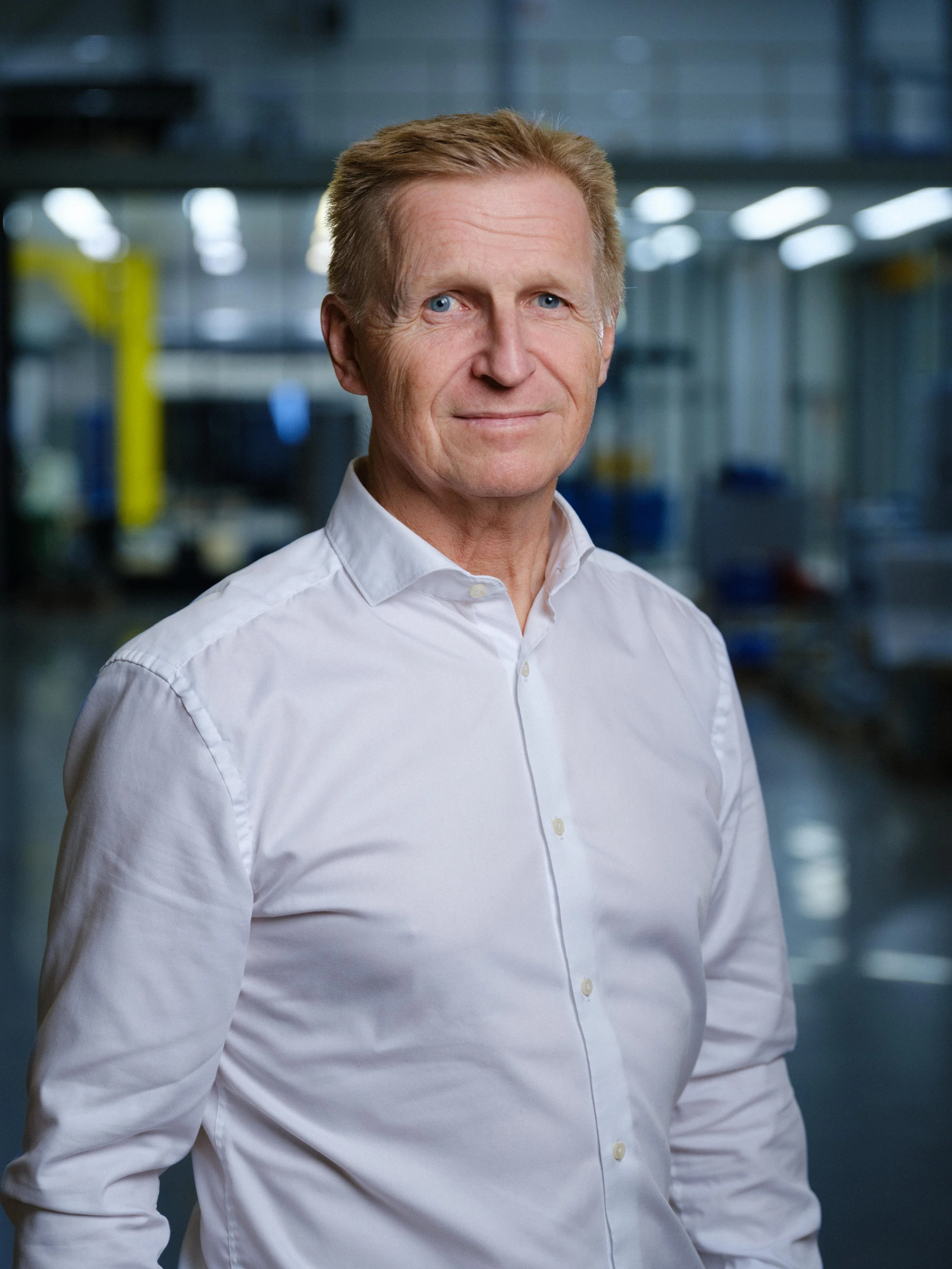 En äldre man med ljusblå ögon och rödfärgat hår, iklädd vit skjorta, står i en industriell miljö.