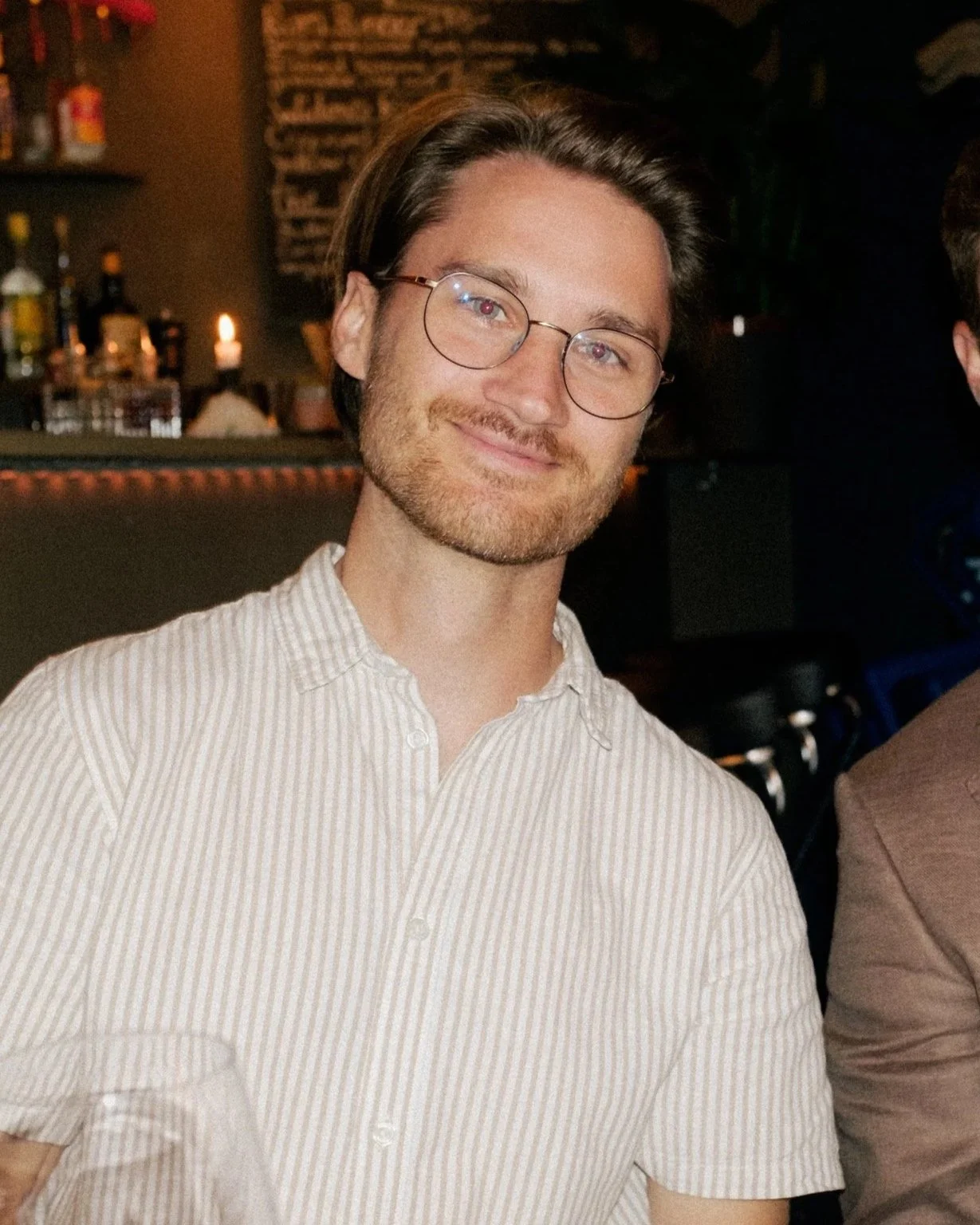 En glad man med glasögon, mörkt hår och skägg, bär en ljus skjorta med ränder, sitter på en restaurang eller bar.