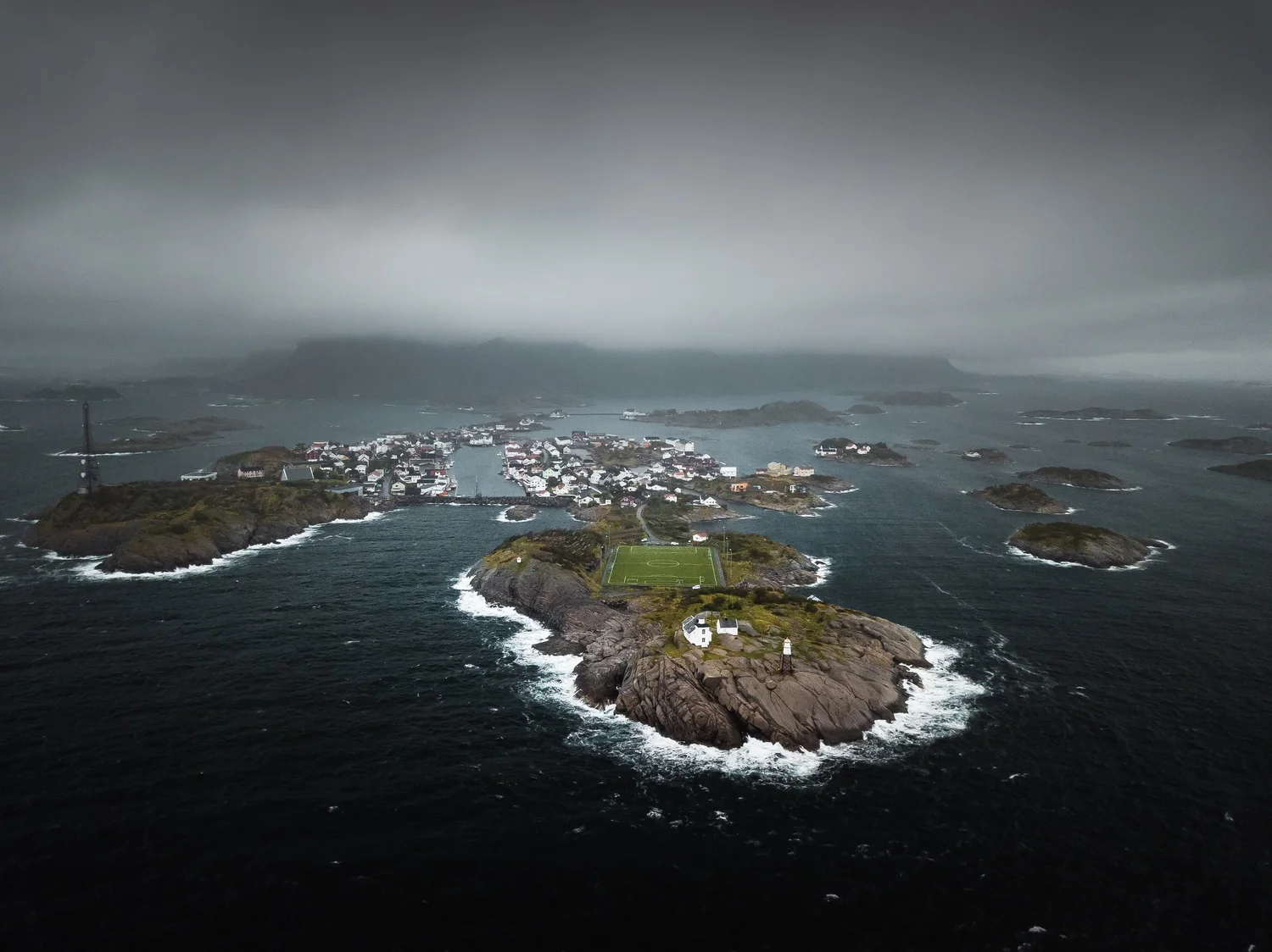 Flygfoto av en kustby med hus och en fotbollsplan mellan berg och hav