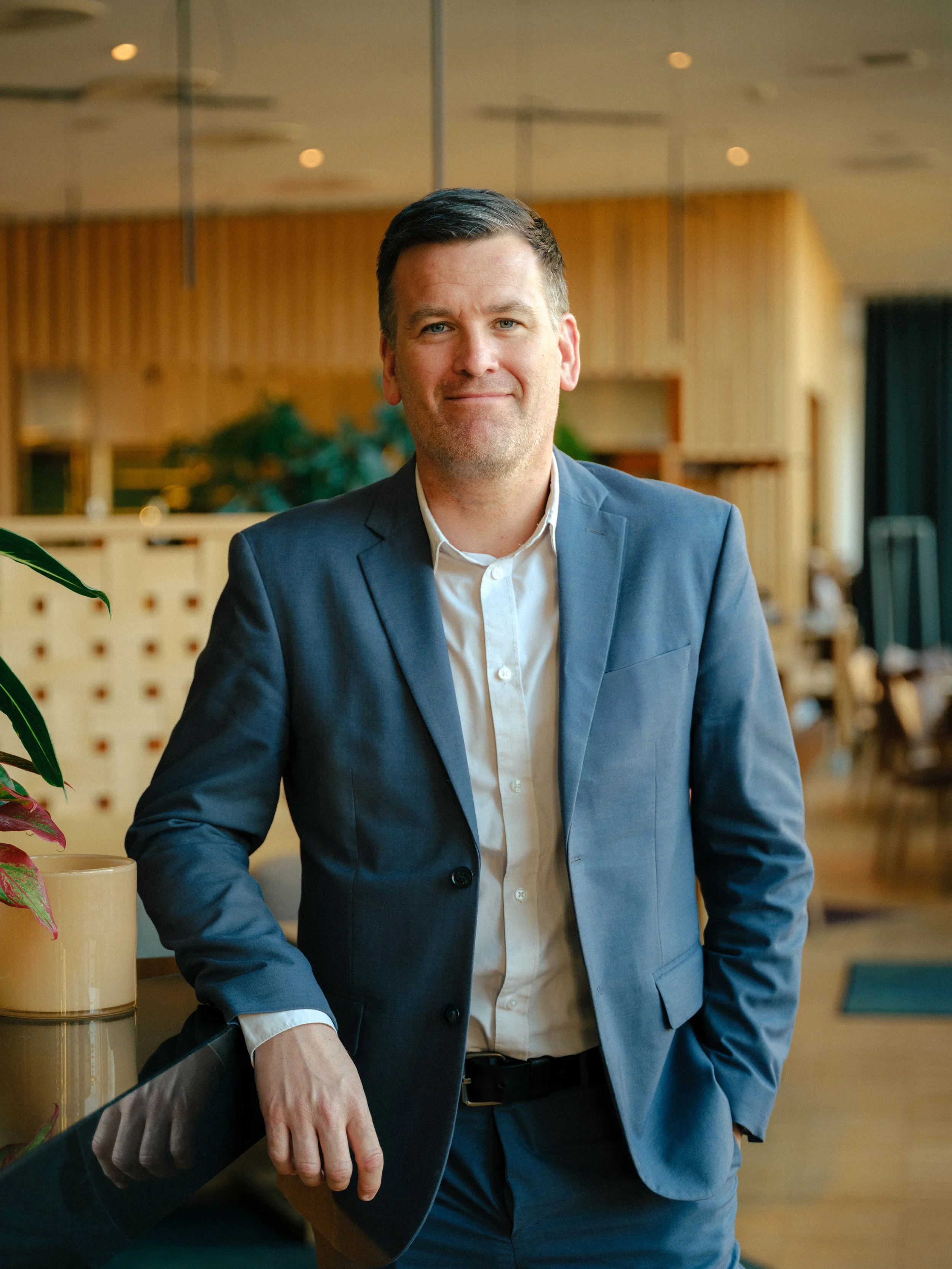 En man i mörkblå kostym står i ett modernt kontorsutrymme, lutad mot en disk med en nöjd uttryck.