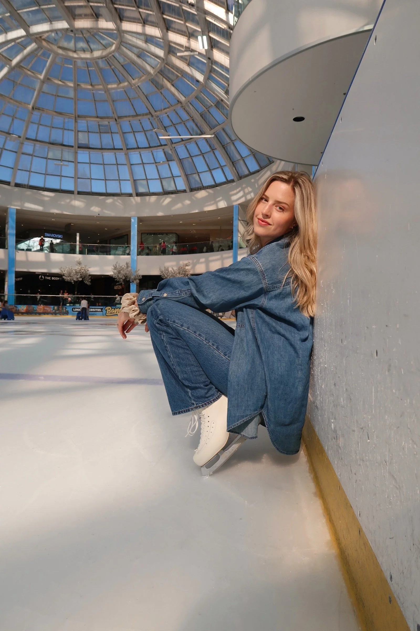Figure skating coach dressed denim seated on the ice with her back to the wall