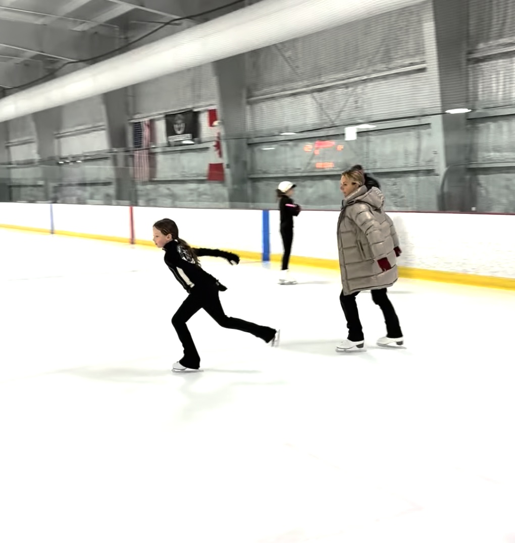 Children ice skating at an indoor rink, with one child in motion and a woman observing, while another girl skates near the boards in the background.