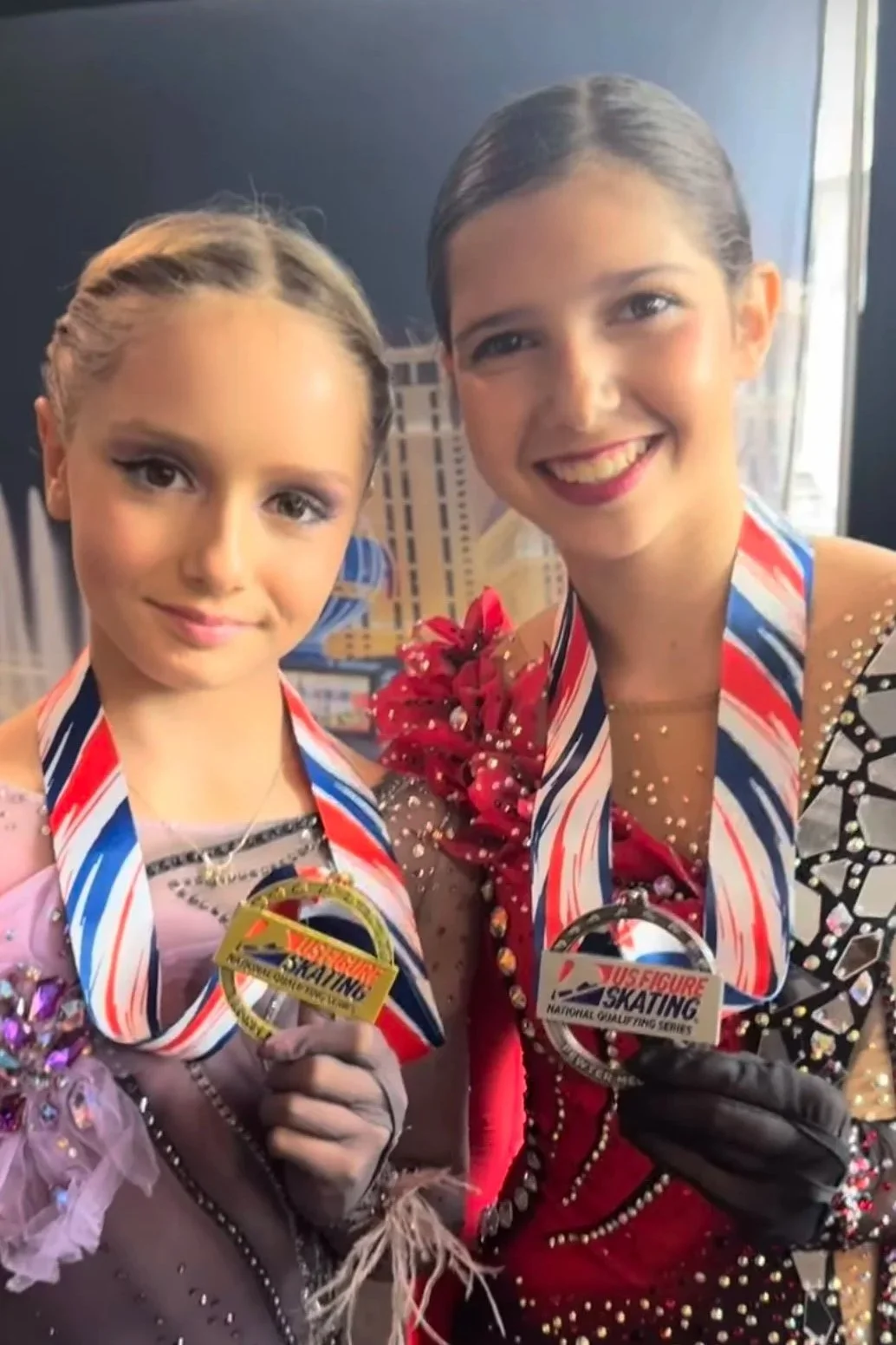 Two young female figure skaters holding medals and smiling, dressed in colorful performance costumes, standing in front of a cityscape backdrop.