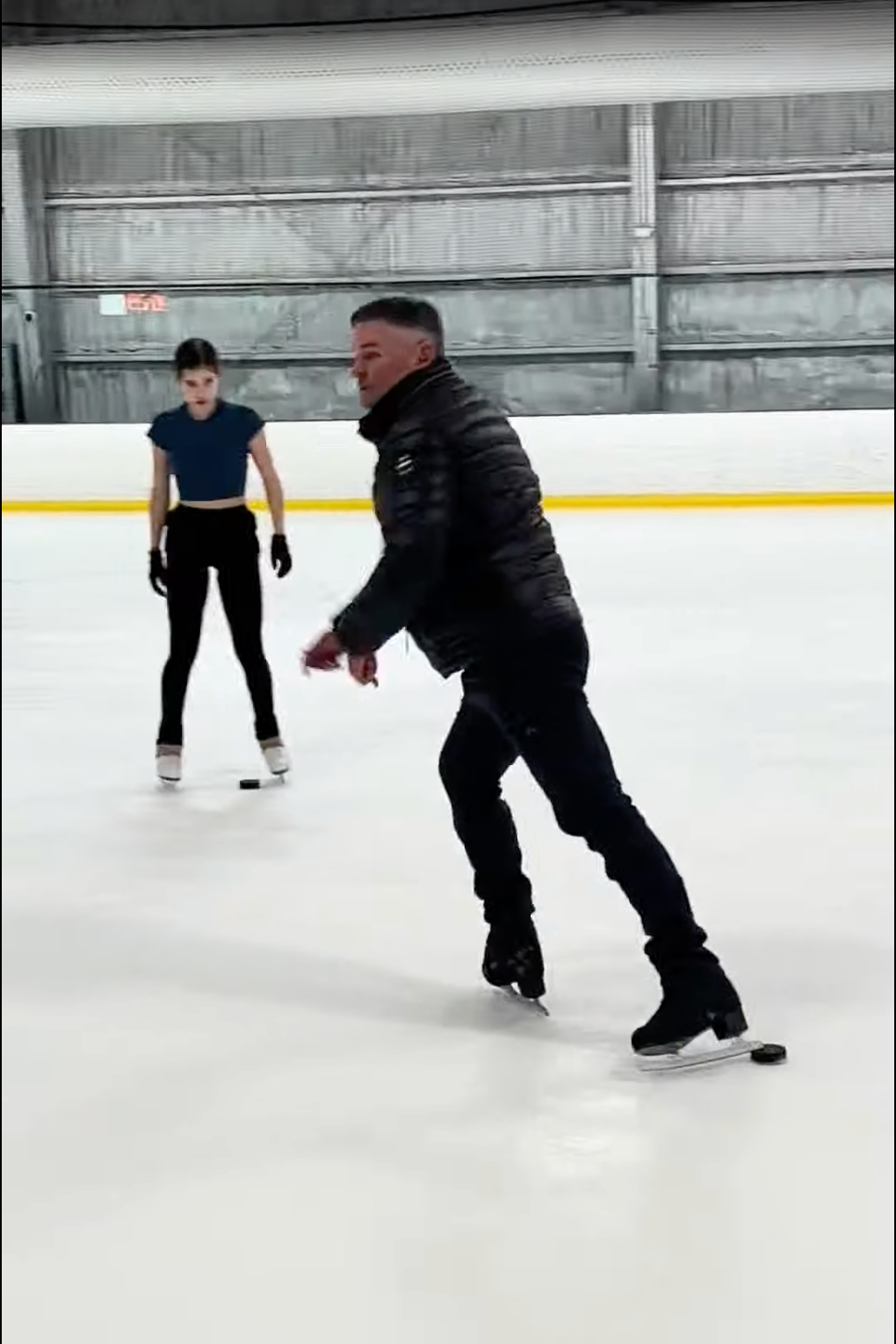 Two people ice skating on an indoor rink, with the person in the foreground performing a skating move and the person in the background watching.