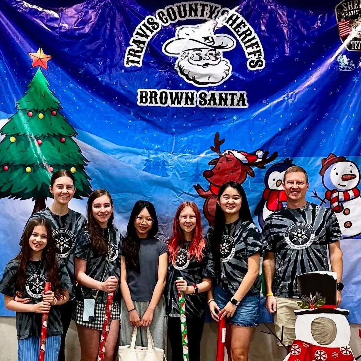 Group of six young people and one adult standing in front of a holiday-themed backdrop with a Christmas tree and cartoon snowmen. They are smiling and holding holiday sticks or presents, celebrating at the Travis County Youth Hockey Brown Santa event.