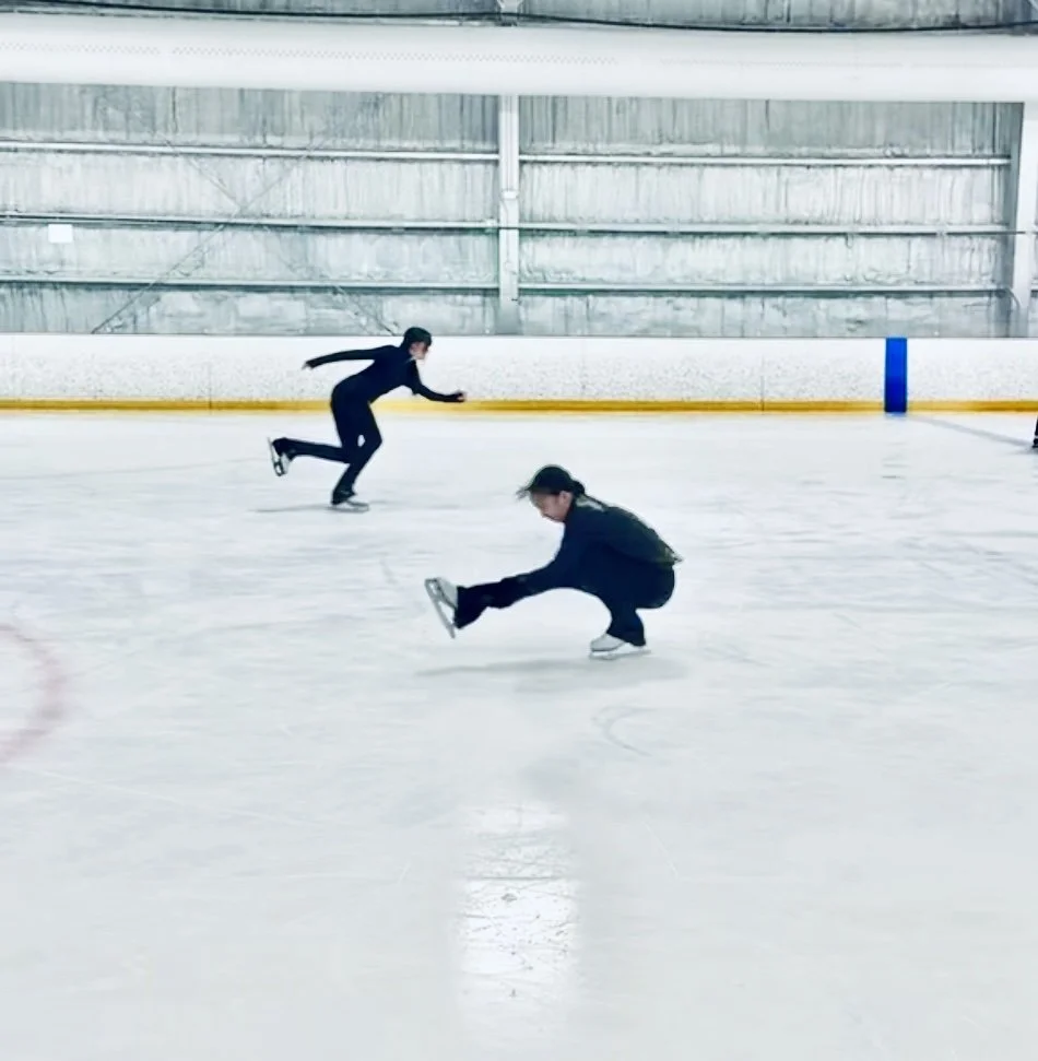 Two women figure skating on an indoor ice rink, one in the background and one in the foreground, both dressed in dark clothing.