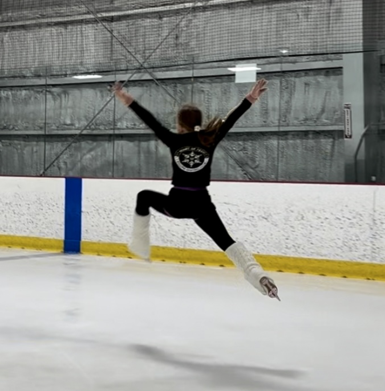 Young girl on ice skates performing a jump at an indoor ice rink, wearing black sportswear and white leg warmers.