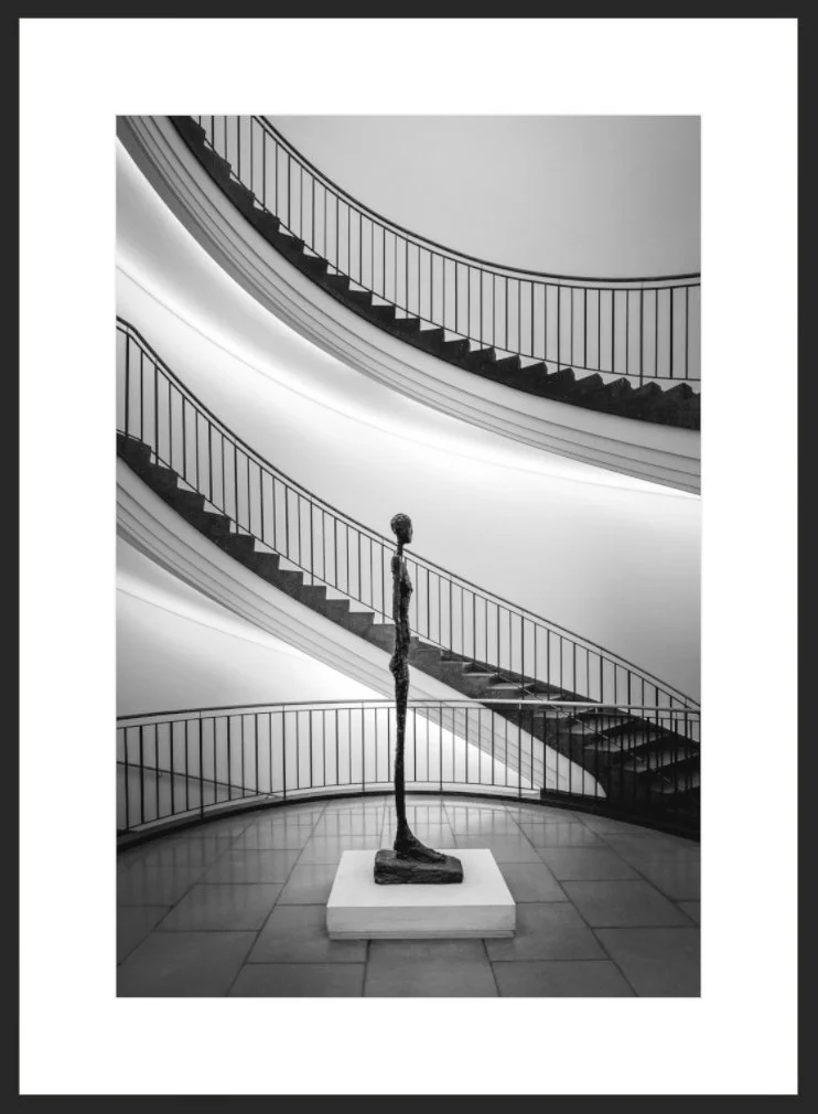Skulptur in einem Kunstmuseum, umgeben von geschwungenen Treppen und weißer Wand in Schwarz-Weiß-Fotografie.