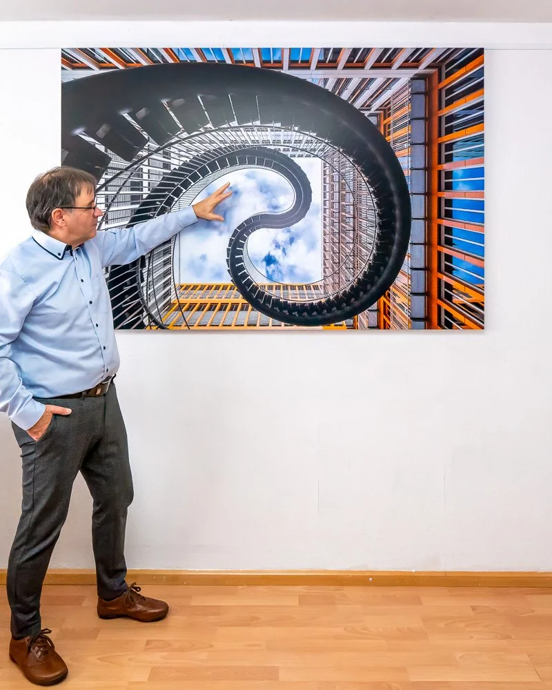 Ein Mann steht vor einem Wandbild, das eine spiralförmige Wendeltreppe zeigt, die nach oben führt und mit Himmel und Wolken im Hintergrund sichtbar ist.