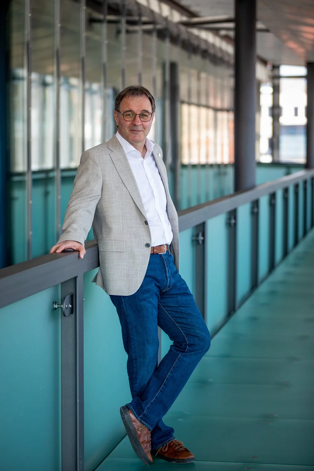 Ein Mann in einem hellen Anzugjacke, weißem Hemd und Jeans lehnt an einer Handläufe in einer modernen, lichtdurchfluteten Halle.