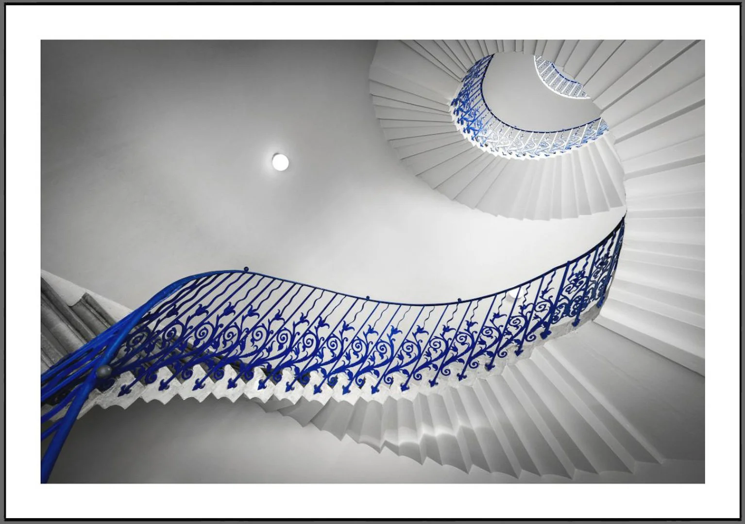 Innenaufnahme einer spiralförmigen Treppe mit blauer verzierter Brüstung, Blick nach oben zur Decke, weiße Wände, Beleuchtungsquelle an der Wand.