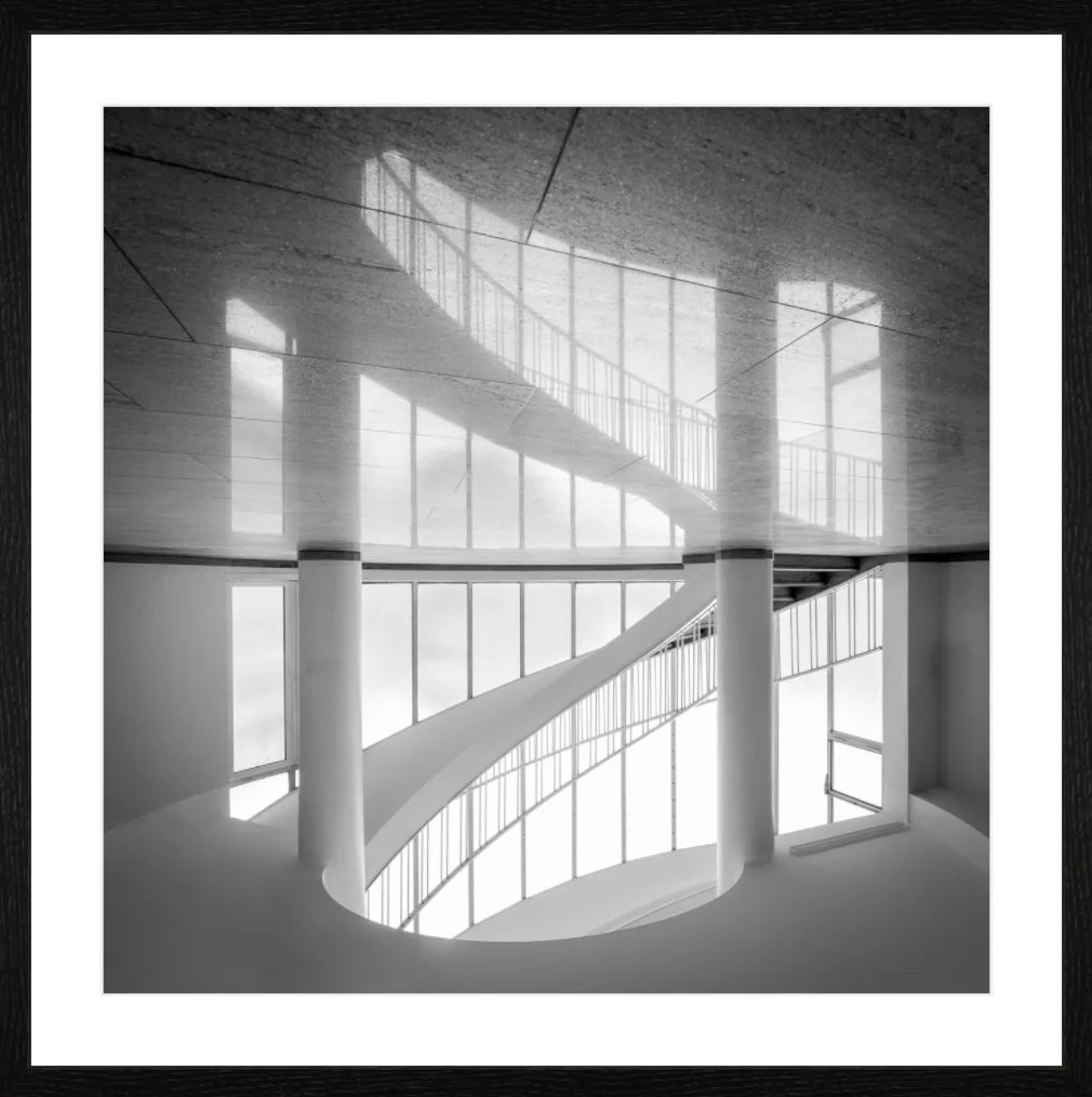 Schwarze und weiße Fotografie eines modernen, spiralförmigen Treppenhauses mit großen Fenstern, die natürliches Licht hereinlassen.