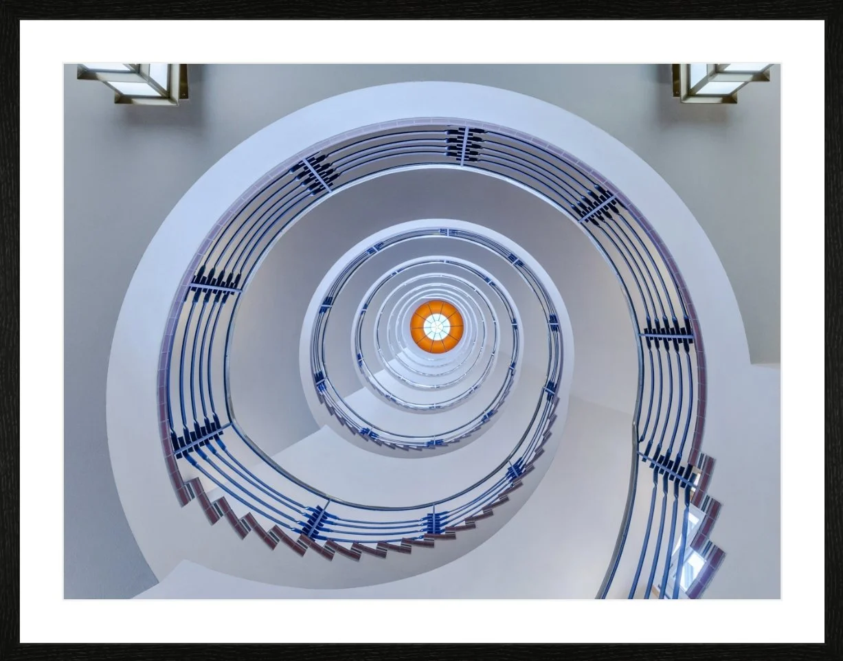 Blick von unten auf eine spiralförmige Treppe mit blauen Geländern, die nach oben führt, mit einer orangefarbenen Lichtquelle am Ende der Treppe.