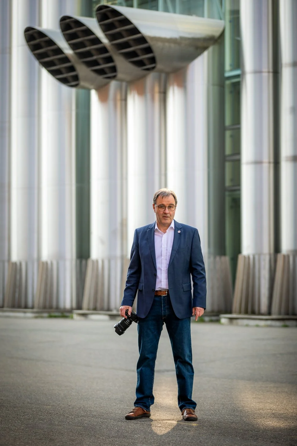 Ein Mann in blauer Jacke und Jeans hält eine Kamera in der Hand, steht vor einem modernen Gebäude mit großen Oberlichtern und metallenen Säulen.