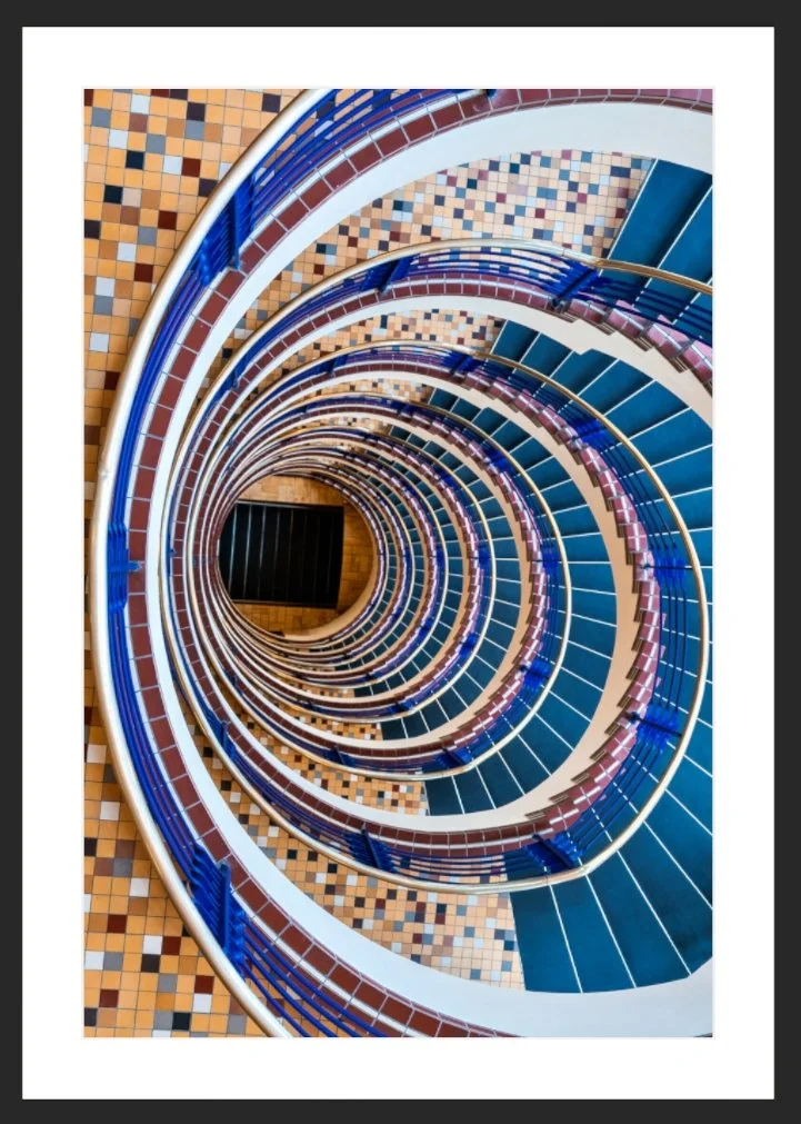 Blick von oben auf eine spiralförmige Treppe in einem Gebäude mit bunten Fliesen in Blau, Braun, Beige und Weiß.